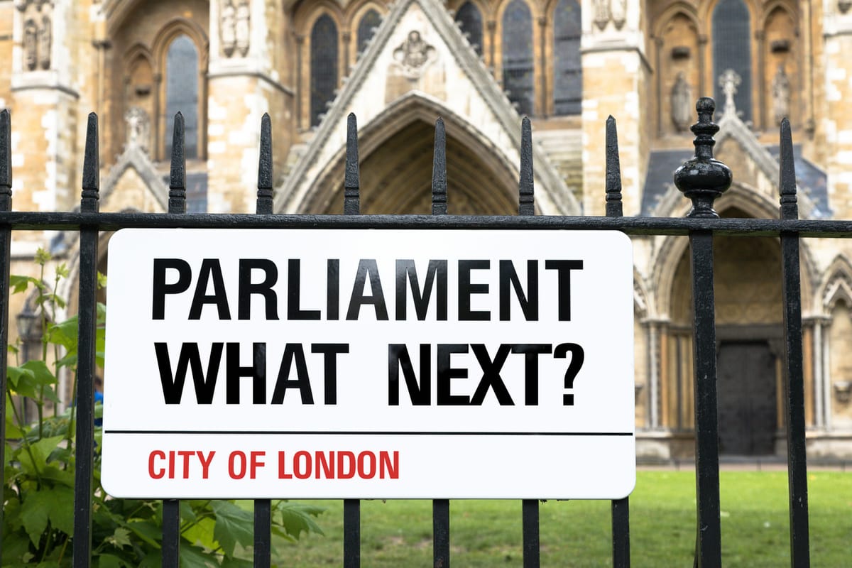Sign in the style of a London Street sign asking Parliament, what next? Black white and red banner against wrought iron railings, with Westminster Abbey in the background.